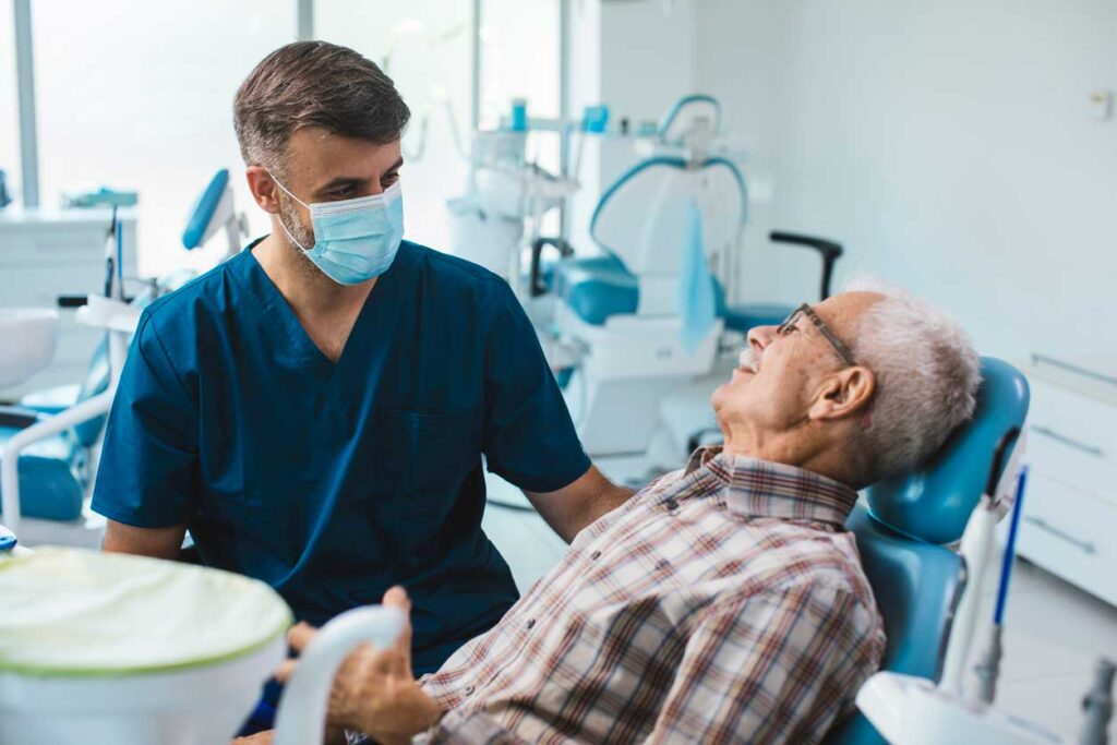 A dental professional in blue scrubs and a face mask, interacting with an elderly patient seated in a dental chair in a clinical setting with dental equipment.